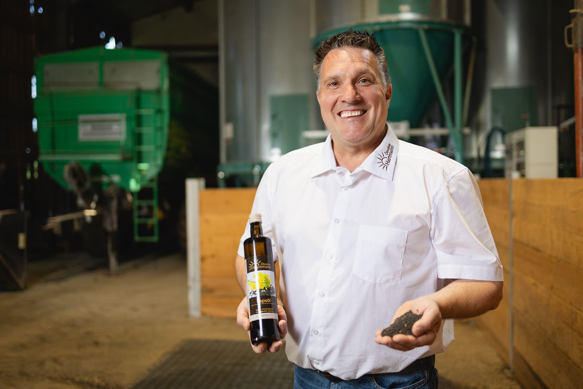 Transparenz und ehrliches Handwerk – direkt aus unserer Ölmühle Marcus Hartmann steht lächelnd in der Produktion der Ölmühle, hält eine Flasche Rapsöl in der einen Hand und präsentiert auf der anderen Hand eine Handvoll Ölsaat.