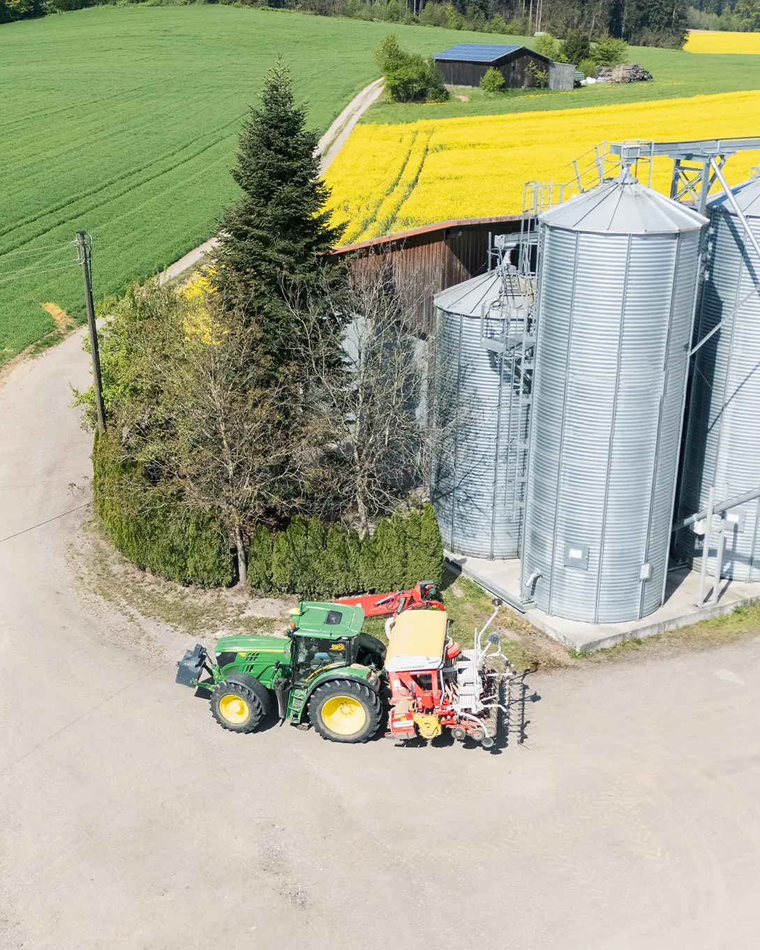 Luftaufnahme der Ölmühle Hartmann mit großen Getreidesilos, einem grünen Traktor samt Sämaschine auf dem Hof und einem blühenden Rapsfeld im Hintergrund.