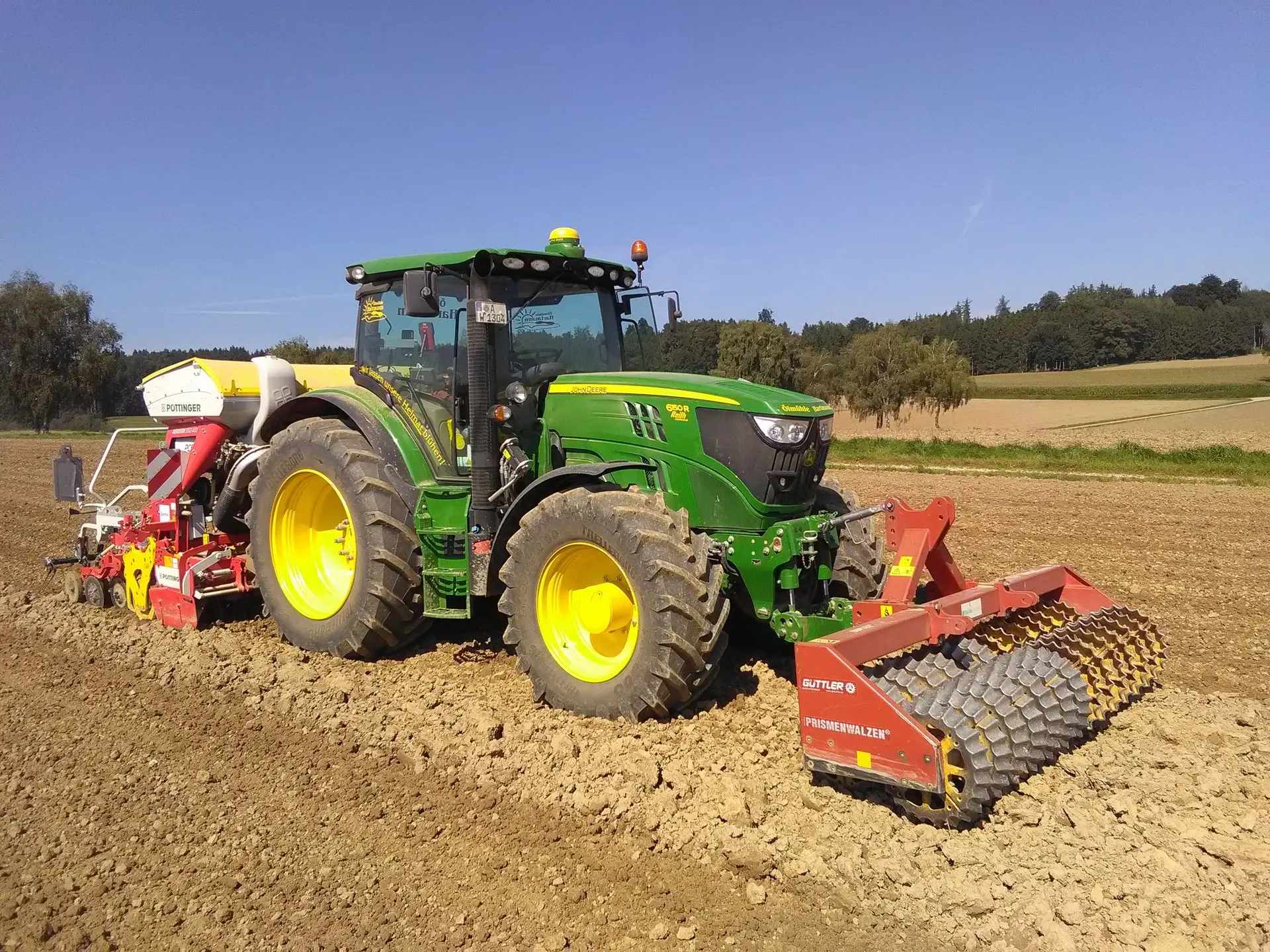 Ein grüner Traktor mit einer roten Sämaschine bei der Feldbearbeitung auf einem frisch gepflügten Acker unter blauem Himmel.