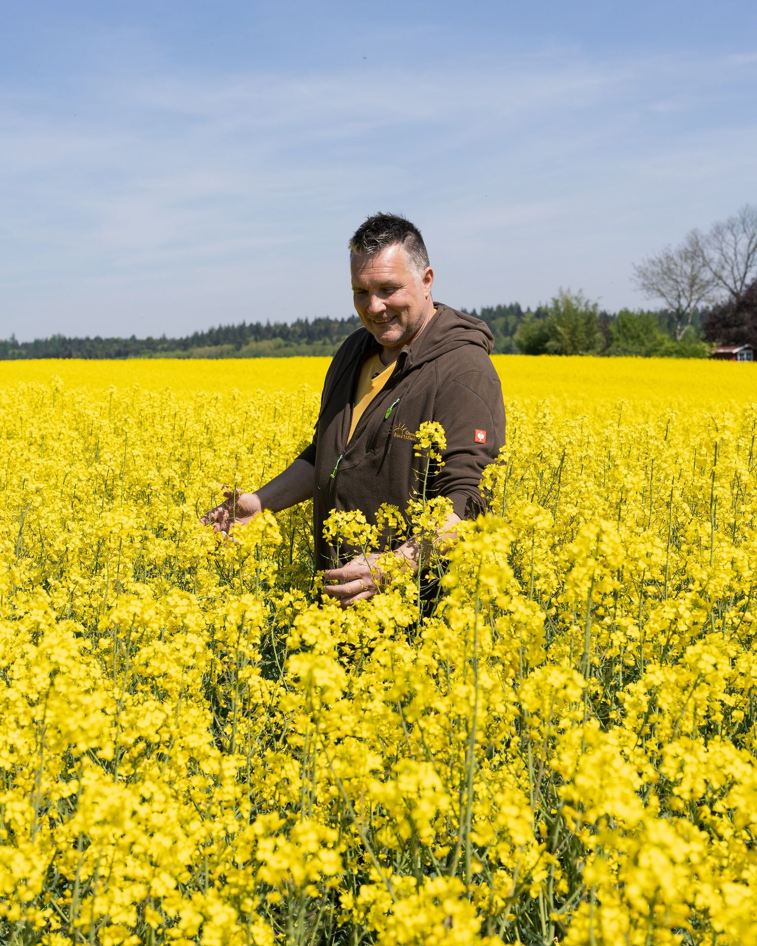 Qualität von Anfang an: Sorgsame Kontrolle unserer Kulturen direkt auf dem Feld Marcus Hartmann steht in einer braunen Kapuzenjacke lächelnd inmitten eines weiten, gelb blühenden Rapsfeldes und begutachtet die Pflanzen.